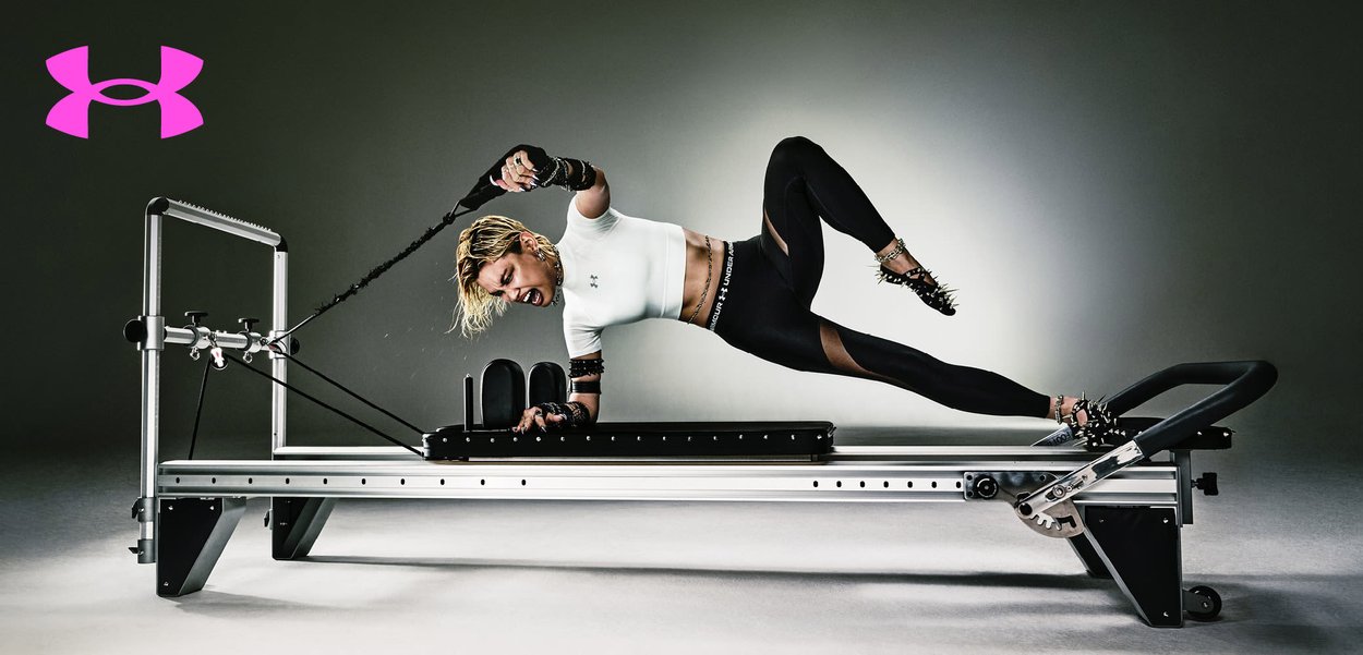 Persoon in Under Armour sportkleding en spikes die oefent op een reformerapparaat met weerstandsbands.