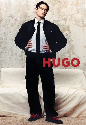 Jeune homme debout, mains sur les hanches, portant une veste bomber noire, une chemise blanche à rayures, une cravate noire, un pantalon noir et des chaussures noires à lacets rouges.