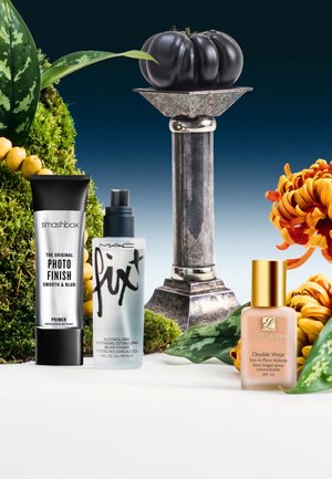 Three cosmetic products: Smashbox primer in a black tube, MAC setting spray in a clear bottle, Estee Lauder foundation in a tan glass bottle. Black pumpkin and metal vase backdrop with green leaves and orange flowers.