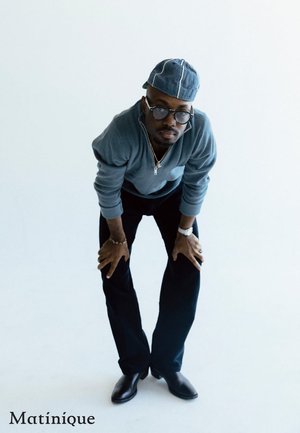 Langärmliger, hellblauer Strickpullover; schwarze, eng anliegende Hose; Jeansmütze; runde Brille; silberner Schmuck; lässige Pose vor einem neutralen Hintergrund.