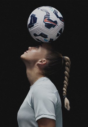 Pallone da calcio Nike equilibrato sulla testa di una persona che indossa una maglietta atletica di colore azzurro chiaro. Sfondo scuro. Capelli acconciati in una coda di cavallo intrecciata.