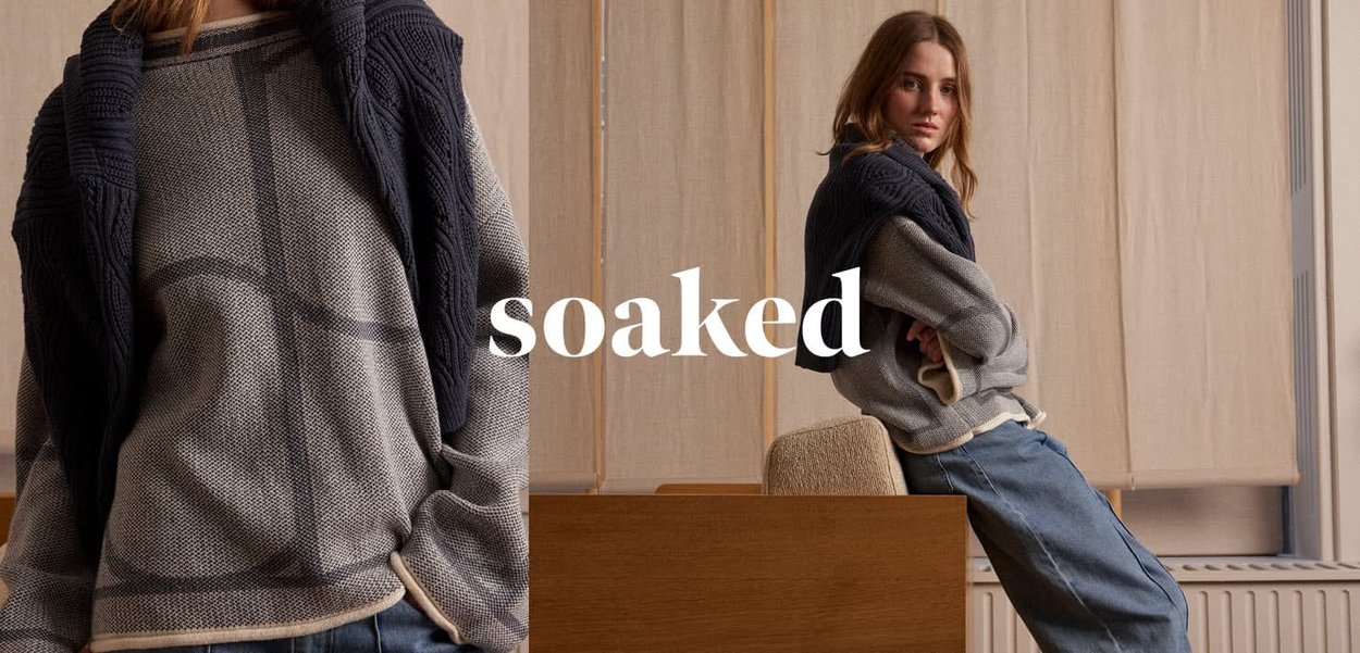 Giovane donna con maglione grigio oversize e jeans blu, maglione blu navy appoggiato sulle spalle, appoggiata a un mobile di legno con testo "bagnato" sovrapposto.