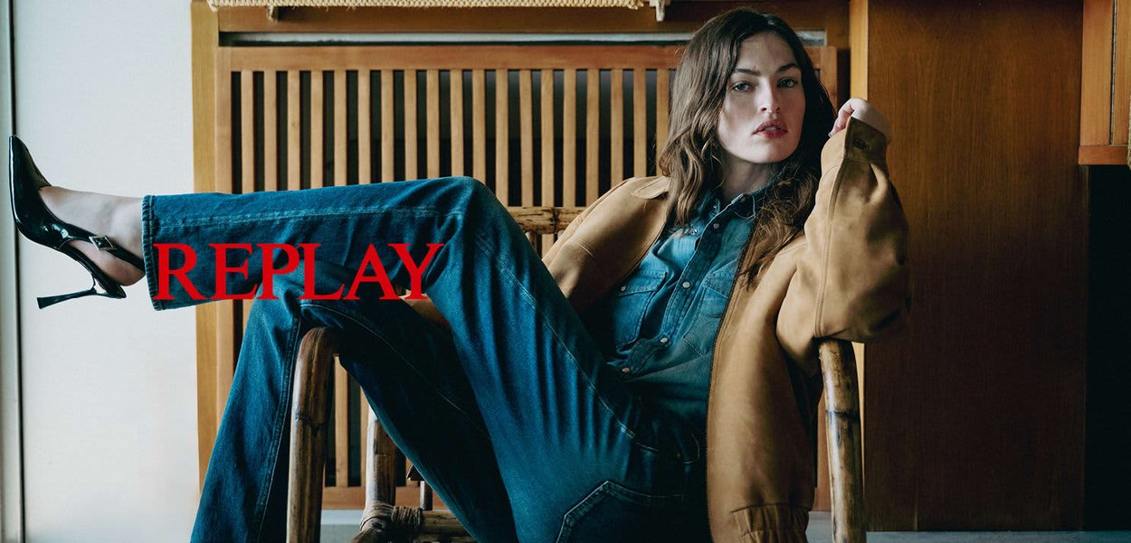 Bruine leren jas, spijkerblouse, donkerblauwe jeans en zwarte hoge hakken, zittend in een bamboestoel tegen een houten achtergrond. Rode tekst "REPLAY."