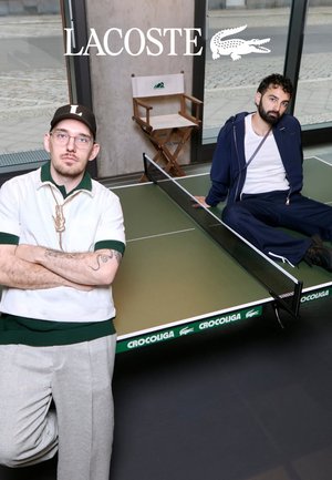 Due uomini posano accanto a un tavolo da ping pong verde Crocoliga al chiuso, uno in piedi con le braccia incrociate, l’altro seduto sul tavolo vicino a una sedia da regista.