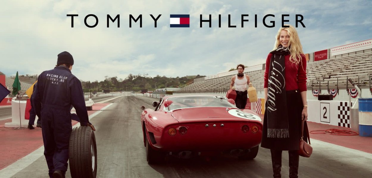 Auto sportiva rossa con una forma elegante e sinuosa, parcheggiata su un circuito. Persone in abbigliamento da corsa e moda sono presenti.