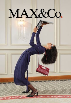 Femme en robe bleu marine et talons, penchée en arrière tenant un ordinateur portable au-dessus d'elle, avec un sac à main rouge sur l'épaule, devant un mur à panneaux et le logo MAX&Co.