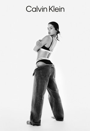 Soutien-gorge de sport noir Calvin Klein avec logo blanc et jean en denim bleu ample, présenté sur un fond blanc. Modèle de profil.