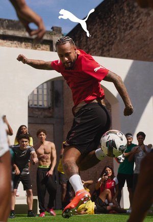 Scarpe da calcio Puma in arancione brillante e giallo; maglia sportiva rossa e pantaloncini neri; giocatore che calcia un pallone su un campo verde.