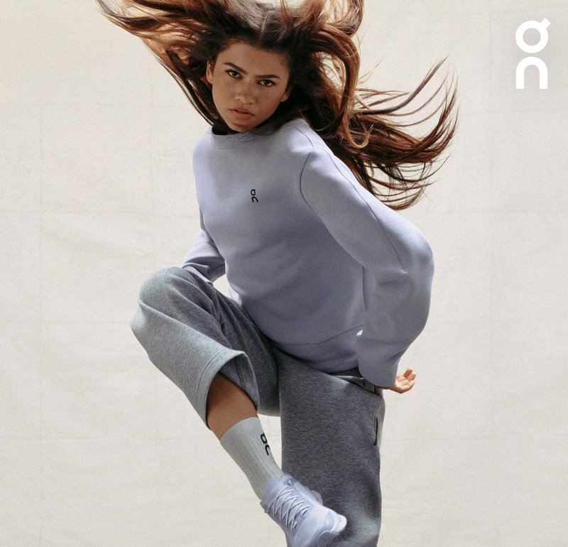 Helllila Sweatshirt mit Logo, kombiniert mit grauen Hosen und weißen Socken. Das Model hat lange, fließende Haare und posiert dynamisch vor einem neutralen Hintergrund.