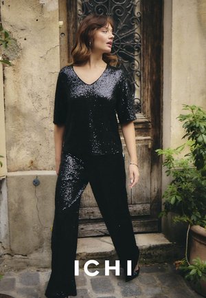 Schwarzes Pailletten-Top mit V-Ausschnitt und kurzen Ärmeln, kombiniert mit passenden Pailletten-Hosen. Glatte Textur, vor einem rustikalen Hintergrund.