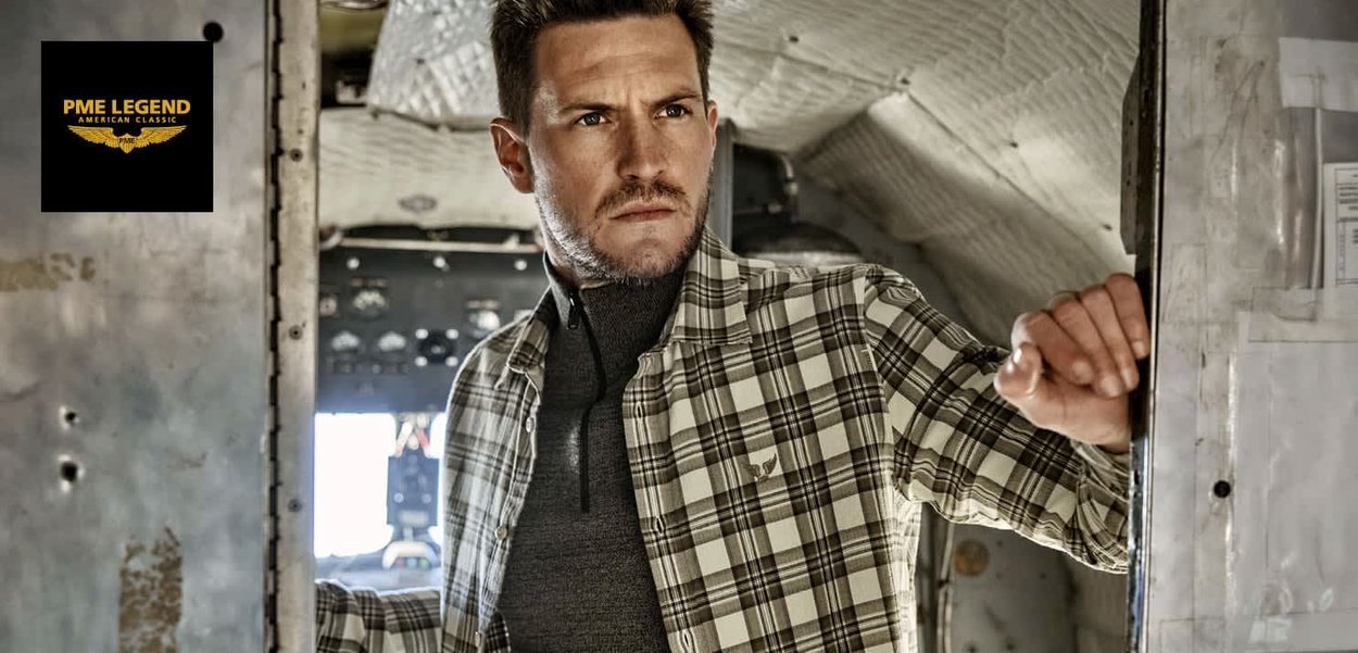 Karoshemd in beige und dunkelgrünen Tönen, kombiniert mit einem schwarzen Reißverschluss-Pullover. Die strukturierte Stoffqualität und die Knopfdetails sind sichtbar.