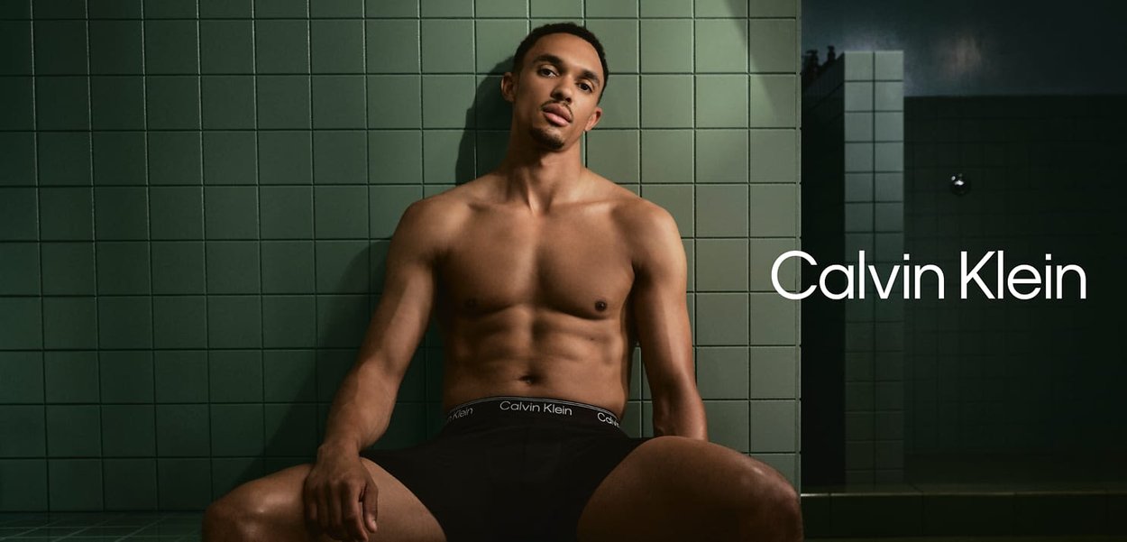 Schwarze Calvin Klein Unterwäsche, die auf einem nackten Oberkörper prangt, vor einem grünen Fliesenhintergrund. Fokus auf dem Stoff und dem kontrastierenden Logo des Waistbands.