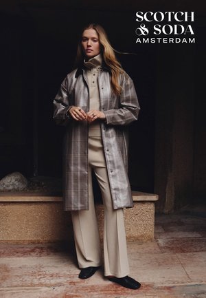 Long manteau à carreaux dans des tons gris porté sur un haut beige et un pantalon à jambes larges. Chaussures noires, bijoux minimalistes, fond texturé avec des éléments en pierre.