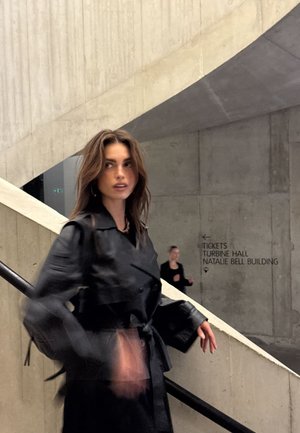 Schwarzer, taillierter Trenchcoat mit strukturierter Oberfläche, übergroßen Ärmeln und breitem Kragen. Getragen von einer Person auf einer Treppe in einem Betonraum.