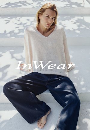Person med lyst hår, der sidder på hvide trin iført en løs cremefarvet sweater og brede mørkeblå bukser med bare fødder synlige.