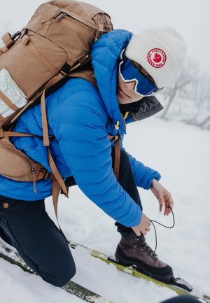 En person knäböjer i snön, iklädd en blå puffjacka, vit keps och solglasögon, med en brun ryggsäck och bruna kängor. Snö och träd i bakgrunden.