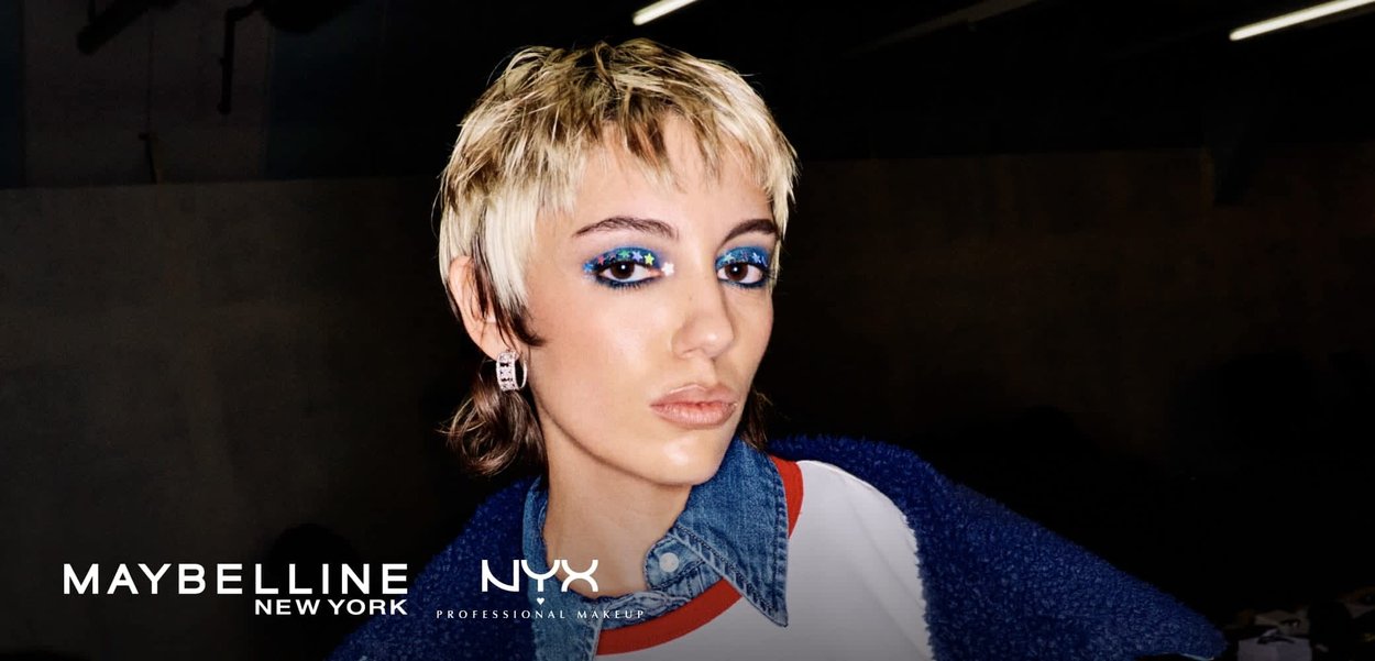 Young person with short blonde hair wears blue star eye makeup, denim shirt, white "COWBOY" shirt, and blue shearling jacket against dark background.