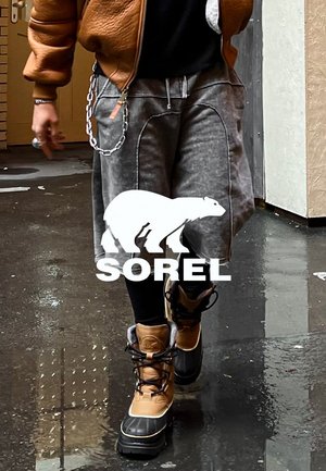 Personne portant des bottes d'hiver imperméables couleur beige et noir, un pantalon gris et une veste marron, marchant sur une surface humide et réfléchissante en extérieur.