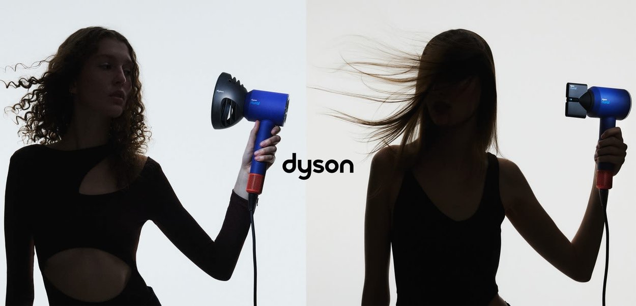 Asciugacapelli Dyson blu con accenti rossi, dotato di un beccuccio curvo. Due modelli tengono il prodotto contro uno sfondo chiaro, mettendo in risalto il design.
