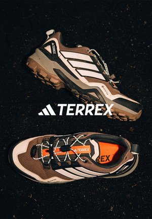 Chaussures de randonnée TERREX marron et blanches avec un tissu texturé, des accents noirs et des détails orange vif ; elles disposent de semelles robustes et de lacets.