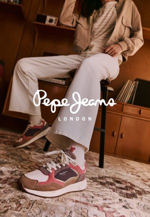 Persona seduta su uno sgabello che indossa pantaloni bianchi e sneakers Pepe Jeans marroni, rosa e viola su un tappeto decorato all'interno.
