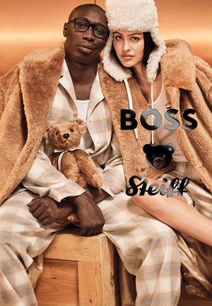 Model-Duo in übergrossen, beigen Kunstfell- und Karo-Loungewear, die einen kuscheligen Teddybär halten. Der Hintergrund zeigt warme metallische Farbtöne.