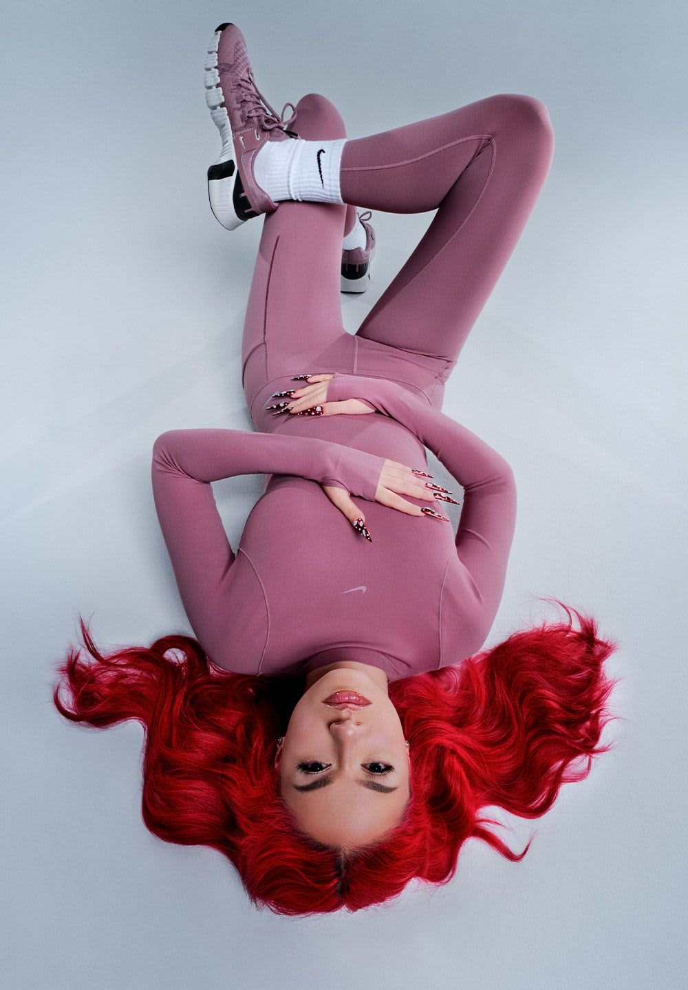 Rosa Sportoutfit, bestehend aus einem enganliegenden Langarmshirt und Leggings, kombiniert mit lila und weißen Turnschuhen. Lange, wellige rote Haare.