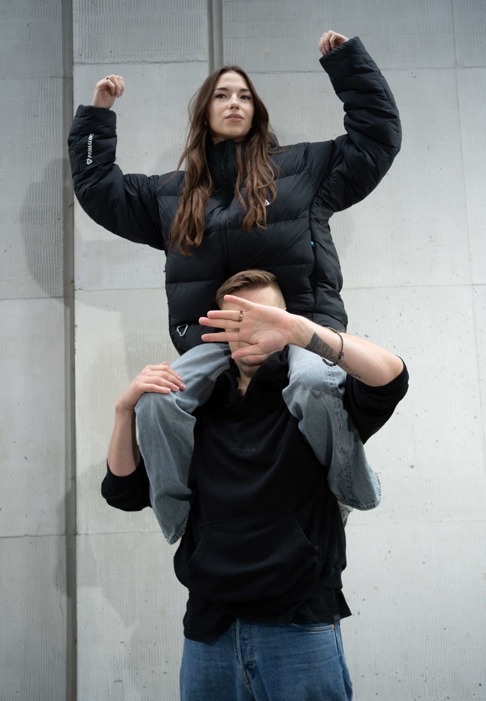 Zwarte pufferjack met een hoge kraag gedragen door de staande persoon. De andere persoon heeft blauwe jeans aan en zit op zijn schouders, met opgetilde armen.