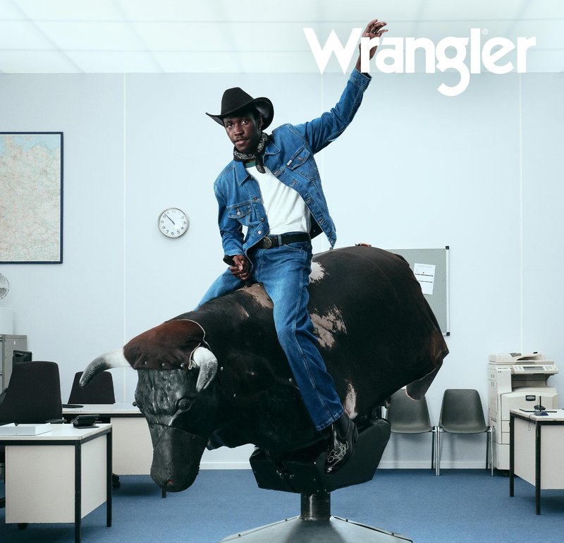 En denim-kledd figur rir på en mekanisk okse med en teksturert, brun og svart overflate. Bakgrunnen har kontormøbler og en klokke.