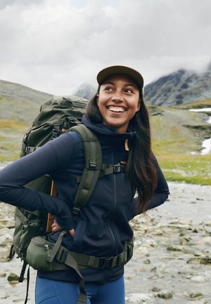 Grön ryggsäck med flera fack, bärs på en marinblå zip-up-jacka. Terrängen är stenig med grön gräs och berg i bakgrunden.