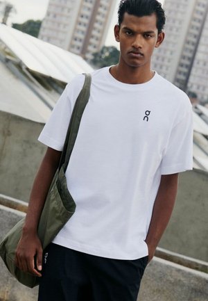 Giovane uomo con una maglietta bianca con un piccolo logo, pantaloni neri e una borsa a tracolla verde oliva, in piedi all'aperto in un'area urbana.