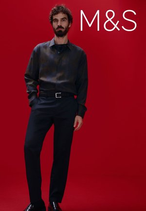 Chemise à carreaux foncés avec un design à boutons associée à un pantalon noir ; le mannequin se tient devant un fond rouge, présentant une tenue moderne.