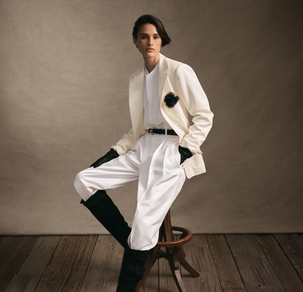 White tailored blazer with a black flower accent, paired with white satin trousers and black leather gloves, set against a neutral backdrop.