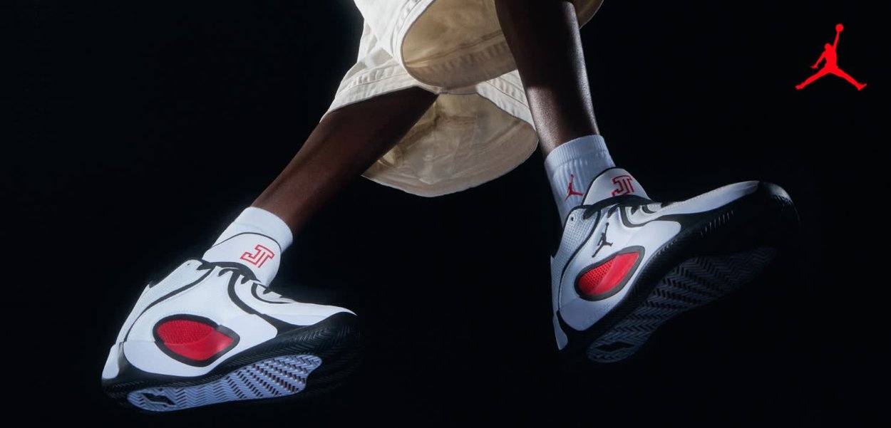 Jambes en short blanc portant des baskets Air Jordan blanches, noires et rouges, sur un fond noir avec un logo Jumpman rouge.