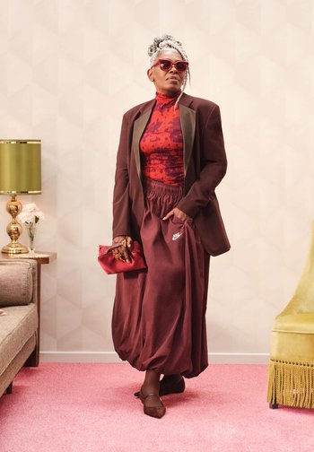 Pantaloni jogger Nike color mogano, dolcevita con schizzi rossi e blazer marrone. Accessori includono occhiali da sole e una pochette rossa. Sfondo con moquette rosa texture.