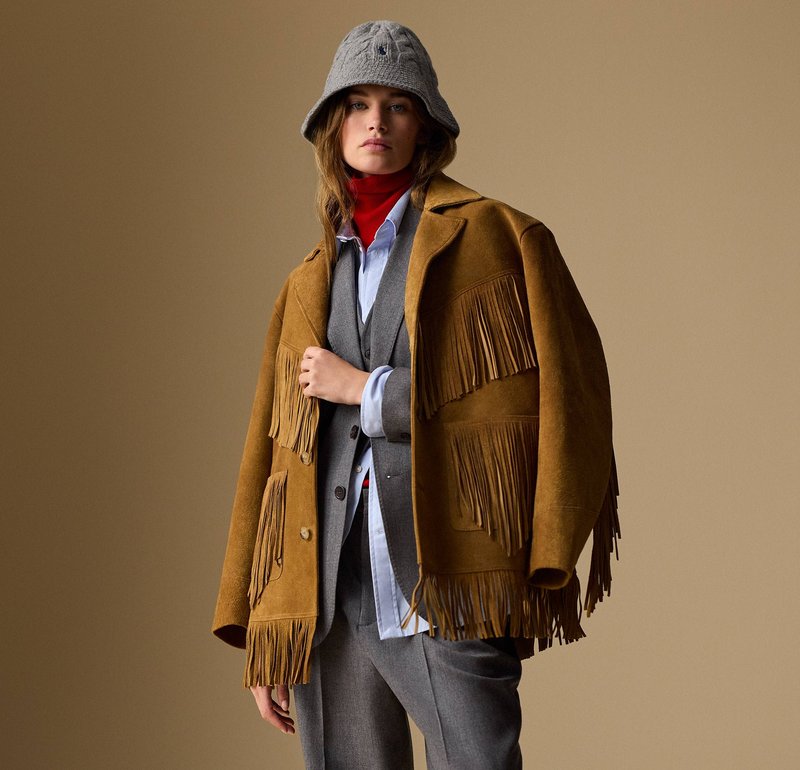 Mujer con un sombrero tipo bucket gris, suéter de cuello alto rojo, camisa azul claro, traje gris y chaqueta de ante marrón con detalles de flecos frente a un fondo liso.