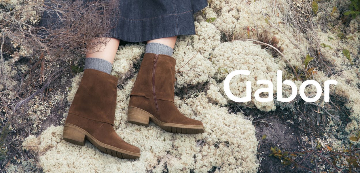 Brown suede ankle boots with a zip closure, low block heel, and rubber sole, paired with grey knitted socks, on mossy ground.
