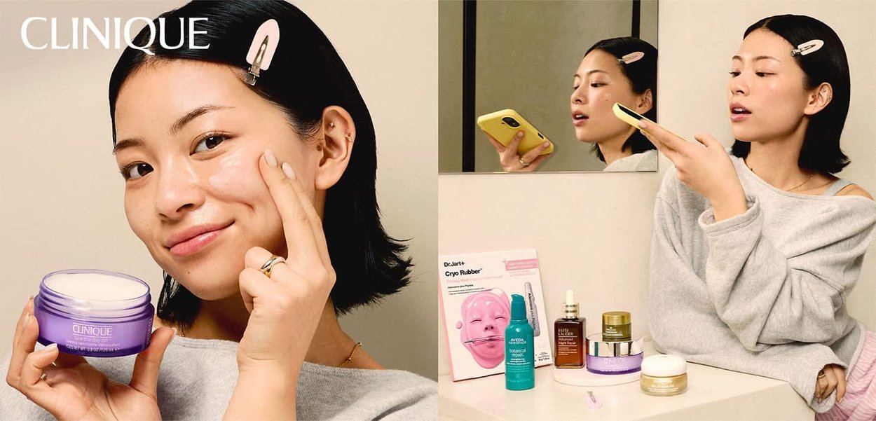 Kvinde med mørkt hår og hårklemme, der påfører Clinique fugtighedscreme, holder en krukke, med flere hudplejeprodukter på bordet og spejl bagved.