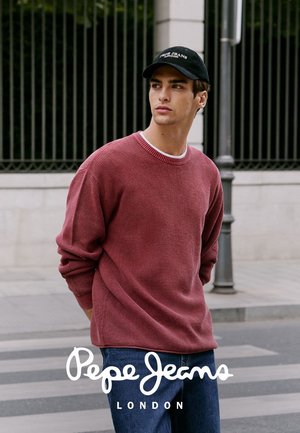Joven con un suéter burdeos, jeans azules y una gorra negra de Pepe Jeans London, de pie en una calle de la ciudad cerca de una barandilla.
