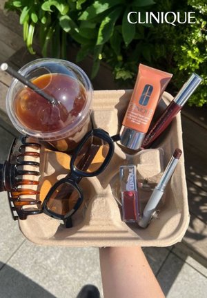 Tray containing sunglasses, iced coffee, and Clinique cosmetics: orange tinted moisturizer, burgundy lip products, and a gray pencil.