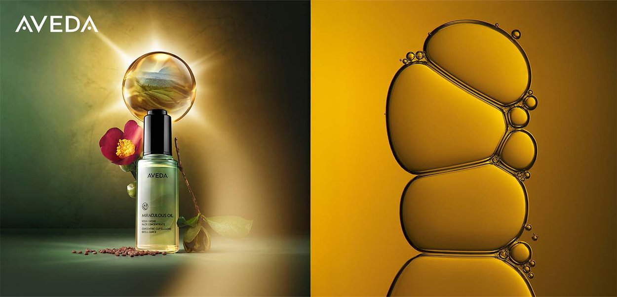 Bottiglia di olio miracolare Aveda, in vetro trasparente con pompa nera, accanto a una foglia e un fiore, su uno sfondo sfumato con gocce d'olio dorato.