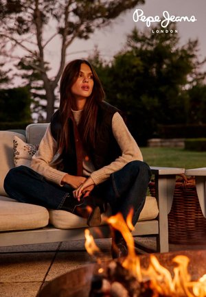 Veste noire portée sur un pull léger, jean bleu foncé et bottes marron. Assis sur un canapé près d'un foyer extérieur, avec de la verdure en arrière-plan.