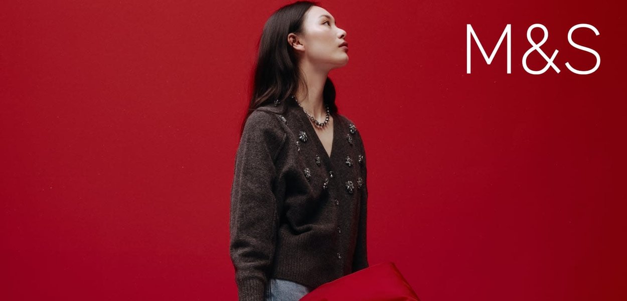 Szary kardigan z kwiatowymi ozdobami, zestawiony z niebieskimi dżinsami. Tło jest jednolicie czerwone. Model trzyma czerwony przedmiot. Widoczne logo M&S.