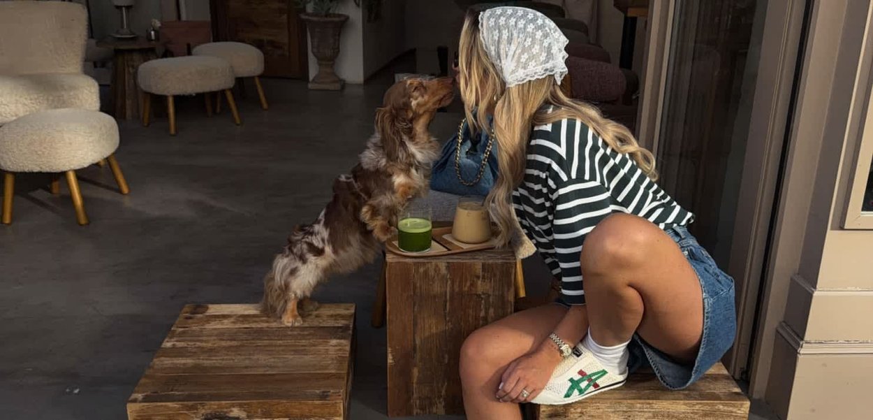 Mujer con camisa de rayas y pantalones cortos vaqueros se arrodilla para besar a un pequeño perro que está sobre un bloque de madera en interiores.