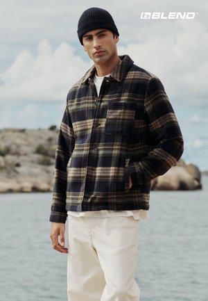 Rutete flanelljakke i marineblå, grønn og krem med brystlomme; kombinert med kremfargede bukser og en svart lue. Naturlig uteområde.