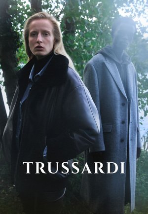 Giacca di pelle con colletto di pelliccia, colore scuro; il modello indossa una camicia di jeans sotto. Cappotto di lana grigia con bottoni, taglio ampio, sullo sfondo.