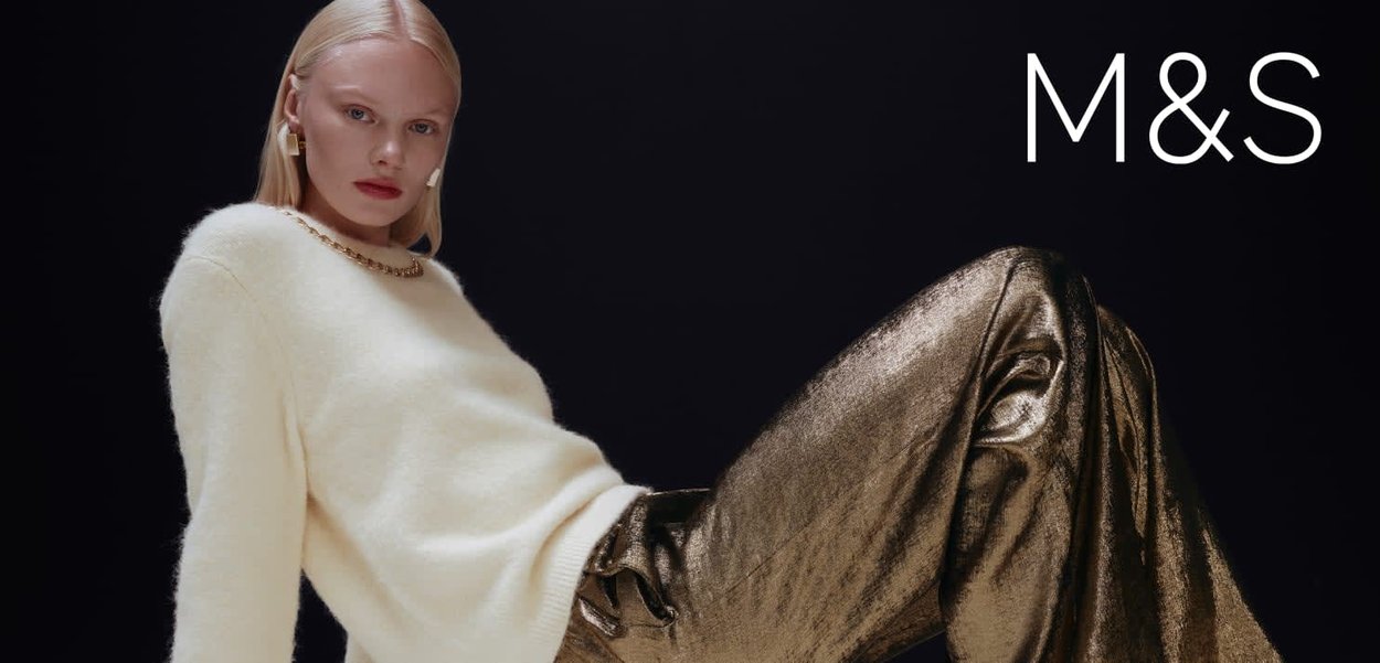 Kremowy sweter z grubym splotem i złotym łańcuszkowym detalem. Błyszczące, metaliczne złote spodnie z szerokimi nogawkami o gładkiej fakturze. Ciemne tło.