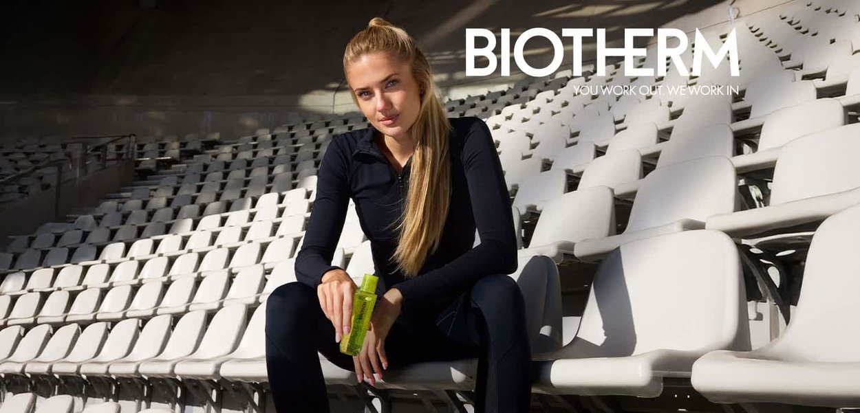 Junge Frau in schwarzer Sportbekleidung sitzt auf Stadionplätzen und hält eine grüne Biotherm-Flasche, Sonnenlicht wirft Schatten.