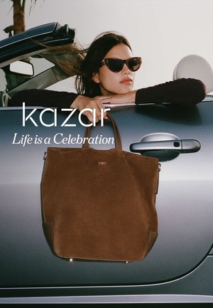 Tote bag in suede marrone con forma strutturata, dotato di accenti in metallo dorato e logo sul davanti, appoggiato contro la portiera di un'auto.