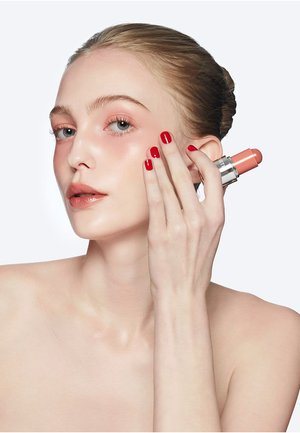 Vrouw met naar achteren gestyled haar brengt perzikkleurige stick-blush aan op de wang, met een gladde huid en rode nagellak op de hand.